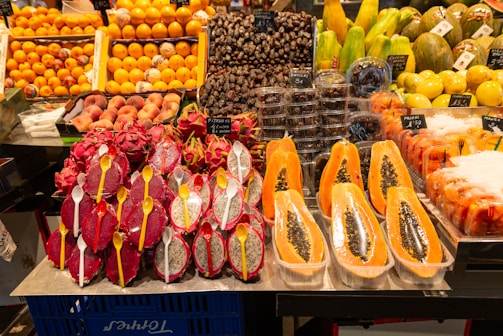 A vibrant display of exotic fruits in a market setting. Front and center are halved papayas, their seeds exposed, alongside vivid red and white dragon fruits with spoons placed inside their halves. Surrounding these are stacks of fresh oranges, dates, peaches, and other tropical fruits like mangoes and passion fruits. Each type is neatly organized with labels displaying their prices.