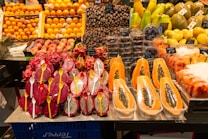 A vibrant display of exotic fruits in a market setting. Front and center are halved papayas, their seeds exposed, alongside vivid red and white dragon fruits with spoons placed inside their halves. Surrounding these are stacks of fresh oranges, dates, peaches, and other tropical fruits like mangoes and passion fruits. Each type is neatly organized with labels displaying their prices.