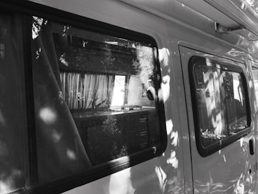 A side view of a van with reflections of trees on the windows. Inside, curtains are visible alongside a kitchenette setup with a stove and counter. The image is in black and white, adding a contrast between the van's exterior and the shadows from surrounding foliage.