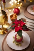 A festive table setting featuring a poinsettia centerpiece placed on a plate with decorative gold elements. The background includes a golden goblet, utensils, and blurred lights, creating a warm and elegant atmosphere.