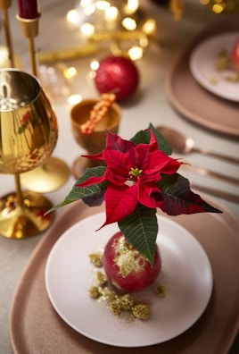 A festive table setting featuring a poinsettia centerpiece placed on a plate with decorative gold elements. The background includes a golden goblet, utensils, and blurred lights, creating a warm and elegant atmosphere.