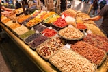 A colorful market display featuring various dried fruits, nuts, and seeds arranged in neat baskets. The assortment includes almonds, pistachios, peanuts, cashews, dried apricots, cranberries, and raisins. Several signs are visible on the baskets, indicating prices or names. The lively scene captures the bustling atmosphere of a market with other shoppers and vendors in the background.