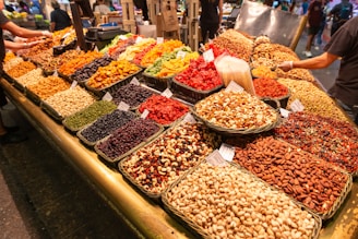 A colorful market display featuring various dried fruits, nuts, and seeds arranged in neat baskets. The assortment includes almonds, pistachios, peanuts, cashews, dried apricots, cranberries, and raisins. Several signs are visible on the baskets, indicating prices or names. The lively scene captures the bustling atmosphere of a market with other shoppers and vendors in the background.
