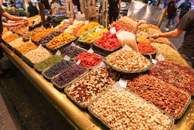 A colorful market display featuring various dried fruits, nuts, and seeds arranged in neat baskets. The assortment includes almonds, pistachios, peanuts, cashews, dried apricots, cranberries, and raisins. Several signs are visible on the baskets, indicating prices or names. The lively scene captures the bustling atmosphere of a market with other shoppers and vendors in the background.