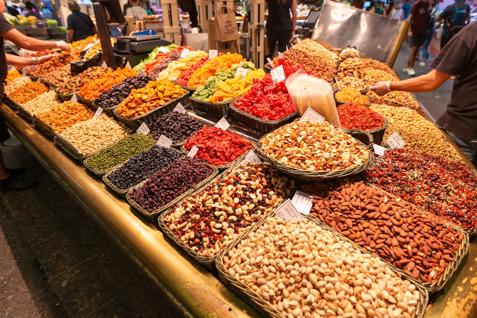 A colorful market display featuring various dried fruits, nuts, and seeds arranged in neat baskets. The assortment includes almonds, pistachios, peanuts, cashews, dried apricots, cranberries, and raisins. Several signs are visible on the baskets, indicating prices or names. The lively scene captures the bustling atmosphere of a market with other shoppers and vendors in the background.