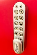 A metallic keypad lock with a vertical arrangement of buttons numbered from one to nine, set against a bright red background. The lock has a modern design with smooth edges and the brand name 'KitLock' displayed prominently at the bottom. The buttons have a beige color with black numbers on them.
