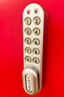A metallic keypad lock with a vertical arrangement of buttons numbered from one to nine, set against a bright red background. The lock has a modern design with smooth edges and the brand name 'KitLock' displayed prominently at the bottom. The buttons have a beige color with black numbers on them.