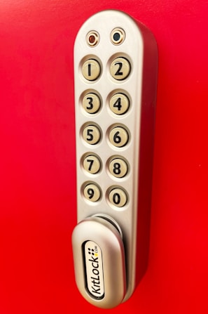 A metallic keypad lock with a vertical arrangement of buttons numbered from one to nine, set against a bright red background. The lock has a modern design with smooth edges and the brand name 'KitLock' displayed prominently at the bottom. The buttons have a beige color with black numbers on them.