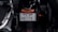 A close-up view of a motorcycle's rear area, focusing on the license plate which includes Chinese characters and the number 46774. The motorcycle is primarily black with orange accents, visible on the rear light and small components. The surrounding area appears dimly lit, highlighting the mechanical elements.