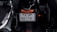 A close-up view of a motorcycle's rear area, focusing on the license plate which includes Chinese characters and the number 46774. The motorcycle is primarily black with orange accents, visible on the rear light and small components. The surrounding area appears dimly lit, highlighting the mechanical elements.