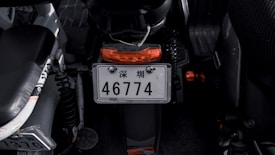 A close-up view of a motorcycle's rear area, focusing on the license plate which includes Chinese characters and the number 46774. The motorcycle is primarily black with orange accents, visible on the rear light and small components. The surrounding area appears dimly lit, highlighting the mechanical elements.