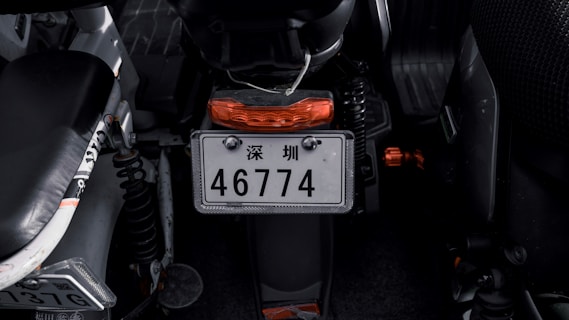 A close-up view of a motorcycle's rear area, focusing on the license plate which includes Chinese characters and the number 46774. The motorcycle is primarily black with orange accents, visible on the rear light and small components. The surrounding area appears dimly lit, highlighting the mechanical elements.