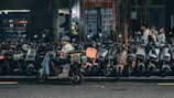 A person wearing a helmet sits on a motor scooter, parked among a large number of similar scooters on the side of the street. The surrounding area is urban, with a backdrop of buildings and storefronts. Other people are walking by, and a tree with drooping branches is visible in the scene.