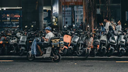 A person wearing a helmet sits on a motor scooter, parked among a large number of similar scooters on the side of the street. The surrounding area is urban, with a backdrop of buildings and storefronts. Other people are walking by, and a tree with drooping branches is visible in the scene.
