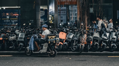 A person wearing a helmet sits on a motor scooter, parked among a large number of similar scooters on the side of the street. The surrounding area is urban, with a backdrop of buildings and storefronts. Other people are walking by, and a tree with drooping branches is visible in the scene.