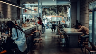 A cozy café with a modern, industrial design featuring high ceilings and large windows. Several people are seated at long tables, using laptops and enjoying their drinks. The café has a bar counter with stools, and a barista is preparing coffee. Outside, there is a view of a street with parked vehicles and a tree-lined background.