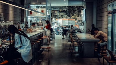 A cozy café with a modern, industrial design featuring high ceilings and large windows. Several people are seated at long tables, using laptops and enjoying their drinks. The café has a bar counter with stools, and a barista is preparing coffee. Outside, there is a view of a street with parked vehicles and a tree-lined background.