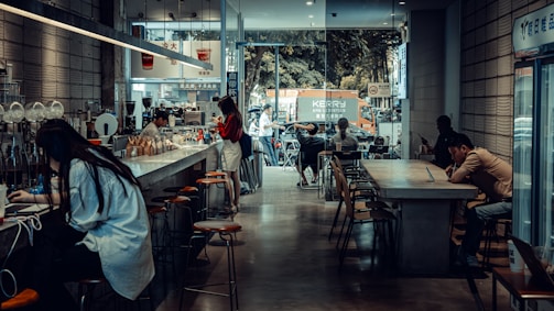 A cozy café with a modern, industrial design featuring high ceilings and large windows. Several people are seated at long tables, using laptops and enjoying their drinks. The café has a bar counter with stools, and a barista is preparing coffee. Outside, there is a view of a street with parked vehicles and a tree-lined background.