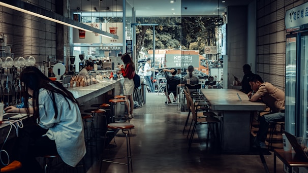 A cozy café with a modern, industrial design featuring high ceilings and large windows. Several people are seated at long tables, using laptops and enjoying their drinks. The café has a bar counter with stools, and a barista is preparing coffee. Outside, there is a view of a street with parked vehicles and a tree-lined background.