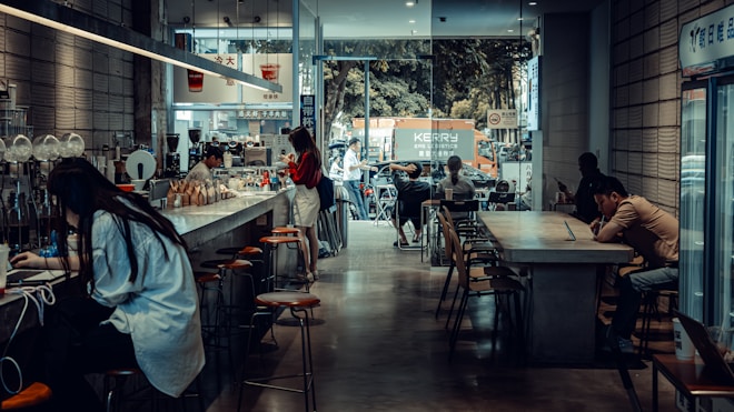 A cozy caf&eacute; with a modern, industrial design featuring high ceilings and large windows. Several people are seated at long tables, using laptops and enjoying their drinks. The caf&eacute; has a bar counter with stools, and a barista is preparing coffee. Outside, there is a view of a street with parked vehicles and a tree-lined background.