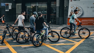 A group of young people with bicycles are gathered in an urban area. They appear to be casually dressed, wearing helmets, and are positioned on a street with prominent yellow grid lines. The backdrop includes a bank entrance and advertisement signs, with an additional child walking in the background.
