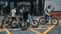 A group of young people with bicycles are gathered in an urban area. They appear to be casually dressed, wearing helmets, and are positioned on a street with prominent yellow grid lines. The backdrop includes a bank entrance and advertisement signs, with an additional child walking in the background.