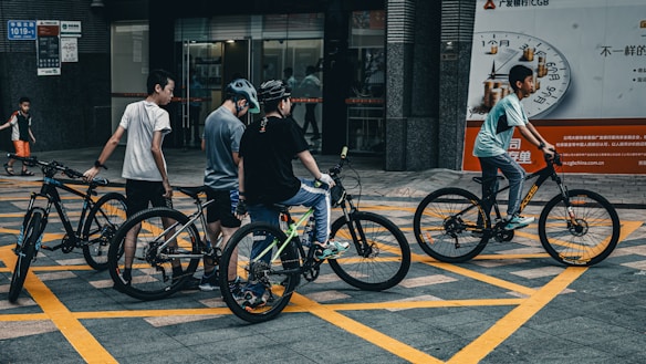 A group of young people with bicycles are gathered in an urban area. They appear to be casually dressed, wearing helmets, and are positioned on a street with prominent yellow grid lines. The backdrop includes a bank entrance and advertisement signs, with an additional child walking in the background.