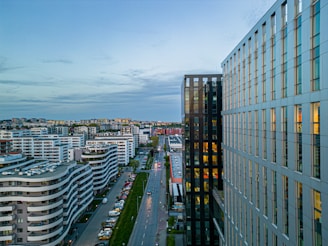 A modern urban landscape featuring high-rise buildings with a mix of glass and concrete facades. The scene includes a long, wide street lined with parked cars and various architectural styles of residential and commercial structures. The sky above shows a gradient of evening light, adding a serene backdrop to the busy urban setting.