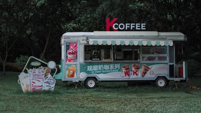 A coffee truck situated in a park-like setting surrounded by lush greenery. The truck features a sign that reads 'K COFFEE' with images of various drinks displayed on its side. Bright and vivid advertisements for ice cream and beverages are visible on the grass next to the truck, promoting colorful and appetizing drinks. The overall scene is serene and inviting, with the food truck standing out against the natural background.
