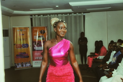 A woman wearing a vibrant pink dress is walking confidently along what appears to be a runway. The room is filled with seated spectators who are observing her. Banners with text and images are seen in the background, suggesting an event or presentation setting.