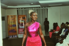 A woman wearing a vibrant pink dress is walking confidently along what appears to be a runway. The room is filled with seated spectators who are observing her. Banners with text and images are seen in the background, suggesting an event or presentation setting.