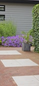 A landscaped garden features a combination of red brick and light gray stone tiles forming a pathway. Lush greenery surrounds the area, including a large leafy plant in a dark gray pot and a vibrant patch of purple flowers against a gray exterior wall. Ivy climbs along one side of the space.