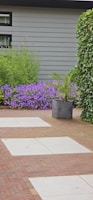 A landscaped garden features a combination of red brick and light gray stone tiles forming a pathway. Lush greenery surrounds the area, including a large leafy plant in a dark gray pot and a vibrant patch of purple flowers against a gray exterior wall. Ivy climbs along one side of the space.