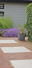 A landscaped garden features a combination of red brick and light gray stone tiles forming a pathway. Lush greenery surrounds the area, including a large leafy plant in a dark gray pot and a vibrant patch of purple flowers against a gray exterior wall. Ivy climbs along one side of the space.