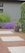 A landscaped garden features a combination of red brick and light gray stone tiles forming a pathway. Lush greenery surrounds the area, including a large leafy plant in a dark gray pot and a vibrant patch of purple flowers against a gray exterior wall. Ivy climbs along one side of the space.