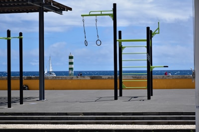 An outdoor gym setup with metal bars and rings is situated on a flat paved surface. Beyond the gym equipment, there is a low yellow wall and a view of the ocean with a sailboat and a green and white striped lighthouse. The sky is partly cloudy.
