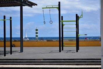 An outdoor gym setup with metal bars and rings is situated on a flat paved surface. Beyond the gym equipment, there is a low yellow wall and a view of the ocean with a sailboat and a green and white striped lighthouse. The sky is partly cloudy.
