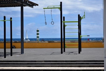 An outdoor gym setup with metal bars and rings is situated on a flat paved surface. Beyond the gym equipment, there is a low yellow wall and a view of the ocean with a sailboat and a green and white striped lighthouse. The sky is partly cloudy.