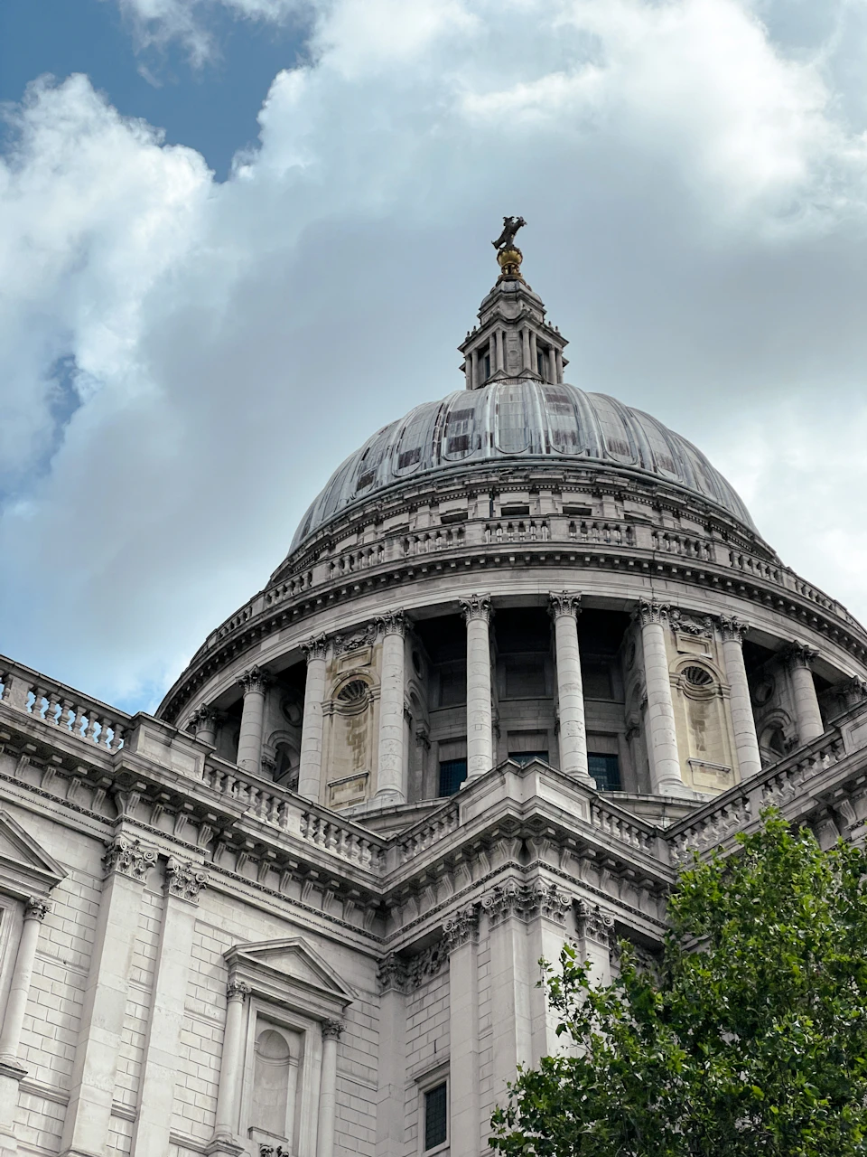 The image shows a majestic white stone building with a dome and columns, standing tall against an overcast sky. The building is adorned with two statues on its roof, one of which is a statue of a lion perched atop the dome. The building features a balcony on the left side, offering a view of the surrounding area. In front of the building, there's a tree that adds a touch of nature to the urban landscape.