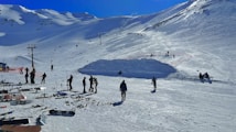 A snowy mountain landscape with numerous skiers and snowboards scattered across a ski resort. Several people are standing and preparing to ski, while others are either walking or standing with their equipment. There are ski lifts and poles set against the backdrop of snow-covered mountains under a clear blue sky.