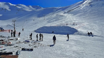 A snowy mountain landscape with numerous skiers and snowboards scattered across a ski resort. Several people are standing and preparing to ski, while others are either walking or standing with their equipment. There are ski lifts and poles set against the backdrop of snow-covered mountains under a clear blue sky.