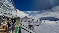A snowy mountain ski resort with people skiing and snowboarding. A modern glass building is on the left, with people sitting and enjoying the view. Ski gear is scattered on the ground, and the clear sky contrasts with the bright snow.