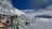 A snowy mountain ski resort with people skiing and snowboarding. A modern glass building is on the left, with people sitting and enjoying the view. Ski gear is scattered on the ground, and the clear sky contrasts with the bright snow.
