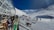A snowy mountain ski resort with people skiing and snowboarding. A modern glass building is on the left, with people sitting and enjoying the view. Ski gear is scattered on the ground, and the clear sky contrasts with the bright snow.