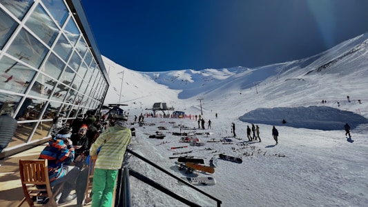 A snowy mountain ski resort with people skiing and snowboarding. A modern glass building is on the left, with people sitting and enjoying the view. Ski gear is scattered on the ground, and the clear sky contrasts with the bright snow.