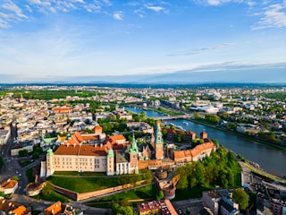 Aerial view of a historic cityscape featuring a prominent castle with red and beige tones, surrounded by greenery and a river flowing alongside. The city is spread out across the landscape, with numerous buildings and roads visible. In the distance, a ferris wheel and modern structures can be seen, adding a touch of contemporary life to the scene.