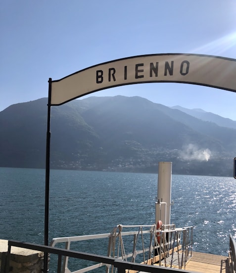 A scenic view of a serene lake with mountains in the background under a clear blue sky. A dock or pier is in the foreground, with a sign reading 'Brienno' arched above the entrance.