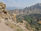 A group of hikers is walking along a narrow, rocky path that runs alongside a steep cliff. The landscape features a winding river nestled in a valley with rugged, forested mountains in the background. The sky is clear with some scattered clouds, and a bird is flying high above the scene.