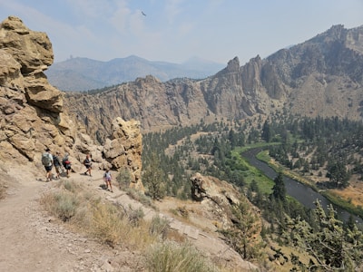 A group of hikers is walking along a narrow, rocky path that runs alongside a steep cliff. The landscape features a winding river nestled in a valley with rugged, forested mountains in the background. The sky is clear with some scattered clouds, and a bird is flying high above the scene.
