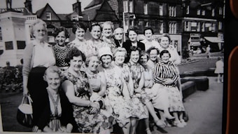 A group of women, dressed in vintage attire, pose together in an outdoor setting. They are seated and standing in front of a backdrop that includes a few buildings and a small park or square. The women are smiling, and the photograph appears to be black and white, suggesting an earlier time period. The area has a quaint, historical feel with visible architectural details.